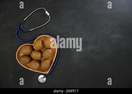 Hohen winkel Foto von vielen leckeren Kiwi in Herzform Holzplatte auf Schwarze Schiefertafel mit medizinische Stethoskop. Stockfoto