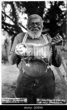 Ein lächelnder, reifer afroamerikanischer Mann steht draußen in einem bewaldeten Gebiet und hält einen toten Possum, 1915. Stockfoto