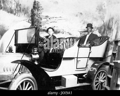 Ganzkörperporträt von zwei Männern in einem Auto, einem jungen kaukasischen Jungen am Steuer, einem afroamerikanischen Mann auf dem Rücksitz, in Anzug und Hut, lächelnde Mimik, 1915. Stockfoto