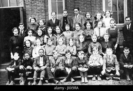 In voller Länge Landschaftsaufnahme von Schulkindern, sitzend und stehend außerhalb des Gebäudes, einige African American, ein Lehrer, Ohio, 1920. Stockfoto