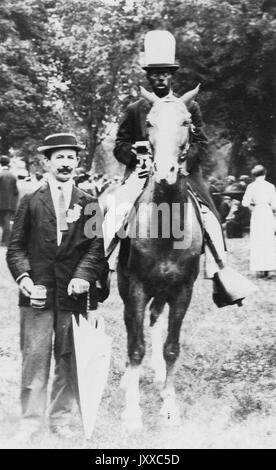 Portrait der afrikanischen amerikanischen Mann reiten und weißer Mann stehend, afrikanische amerikanische Mann mit dunklen Jacke, Hose und Hut, weißen Mann mit dunklen Jacke, dunkle Krawatte, dunkle Hose und Hut, weißen Mann, der Regenschirm, sowohl außerhalb, neutrale Ausdrücke, 1920. Stockfoto