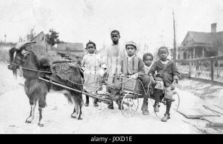 Ein junger afroamerikanischer Junge und Mädchen mit neutralem Ausdruck stehen neben einem offenen Wagen, in dem drei junge afroamerikanische Jungen mit neutralem Ausdruck sitzen, der Wagen, der von einer Ziege getragen wird, auf einer Feldstraße in einer ländlichen amerikanischen Stadt, möglicherweise Savanna, Georgia, 1915. Stockfoto