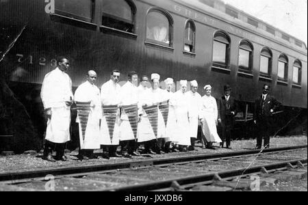 Eine Gruppe amerikanischer Arbeiter, einige in Köchenkleidung und mit Wimpeln, einige formell in dreiteiligen Anzügen gekleidet, stehen draußen an Zuggleisen vor dem Wagen mit der Aufschrift "Great Northern [Railroad]", 1915. Stockfoto