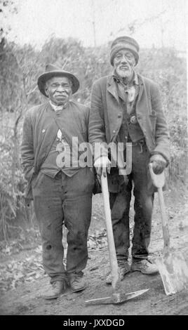 In voller Länge stehend Porträt von zwei reifen afroamerikanischen Arbeiter mit neutralen Ausdrücke stehen in einem gerodeten Waldgebiet mit einem Pickax und einer Schaufel, 1920. Stockfoto