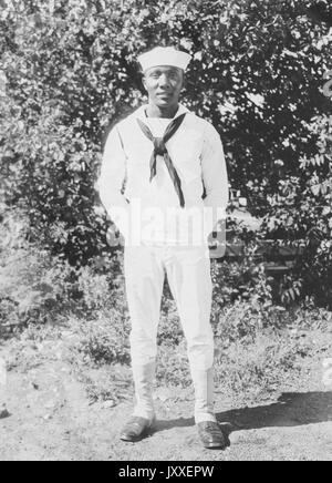 Porträt eines African American US Navy Sailor vor Gebüsch stehend, Hände sind hinter seinem Rücken versteckt und er trägt eine helle Sailor Uniform, 1920. Stockfoto