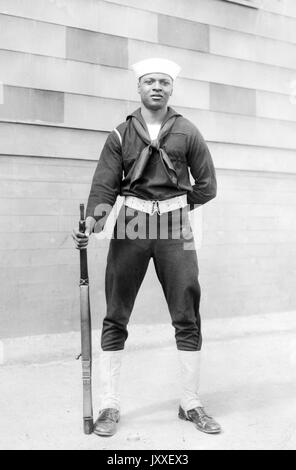 Porträt eines afroamerikanischen US Navy Sailors vor der Wand eines Gebäudes, eine Waffe hinter dem Rücken und die andere hält eine Feuerwaffe an seiner Seite, er trägt einen dunklen Sailor Anzug und einen hellen Sailor Hut, 1920. Stockfoto