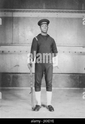 Portrait einer afrikanischen amerikanischen US Navy Sailor stand vor einem großen Schiff, er trägt einen dunklen Sailor Uniform, einen Arm hinter den Rücken und die andere ist im Besitz einer Feuerwaffe durch seine Seite, 1920. Stockfoto