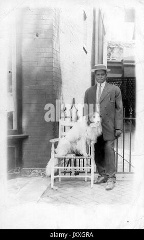 Ganzlanges Standbild des jungen afroamerikanischen Mannes, in dunklem Anzug, hellem Hemd, Hut und Krawatte, neben hellem Hund stehend mit dunklem Sport auf Stuhl sitzend, neutraler Ausdruck, 1930. Stockfoto