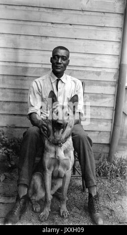 Ganzkörperportrait des reifen afroamerikanischen Mannes, trägt helles Hemd und dunkle Hose, sitzt draußen auf Stuhl vor dem Haus mit Schäferhund sitzen, neutraler Ausdruck, 1925. Stockfoto