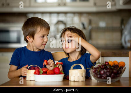 Zwei süße Kinder, junge Brüder essen frisches Obst zu Hause, Erdbeeren, Kirschen, Himbeeren, Heidelbeeren Stockfoto