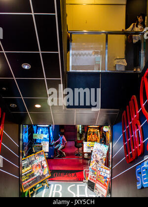 Eine junge Frau, die das Rauchen einer Zigarette, blickte von ihrem Telefon, während sich unter einen Mann spielt ein Video Spiel, Club Sega, Shibuya, Tokio, Japan Stockfoto