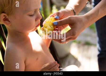 Kaukasische Toddler Boy ohne Hemd auf, sitzt, während seine Mutter ihm zeigt, wie man Essen Mais am Kolben Stockfoto