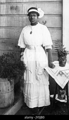 Porträt einer afroamerikanischen Frau, sie trägt ein langes, helles Kleid und eine große Halskette, sie trägt auch ein helles Stirnband mit einer großen Schleife oder Dekoration darin, Links von ihr befindet sich ein Fass Strauchwerk und links von ihr ein runder Holztisch, der runde Holztisch hat zwei Ebenen, auf jeder Ebene ist ein Blumentopf, auf dem runden Tisch liegt sie ihre linke Hand, 1915. Stockfoto