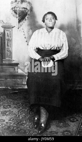 Ganzkörperportrait der afroamerikanischen Frau, vor einer Kulisse sitzend, mit gefalteten Händen auf dem Schoß, in dunklem Kleid und gestreifter Bluse, mit ernsthaftem Gesichtsausdruck, 1920. Stockfoto