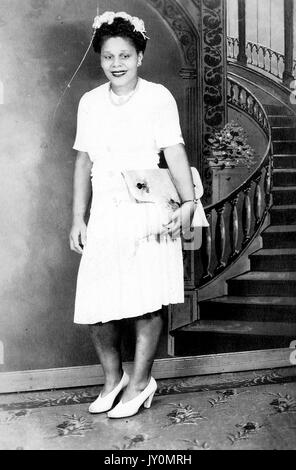 Ganzkörperportrait einer afroamerikanischen Frau, die ein weißes Kleid und weiße Schuhe trägt, ihre Handtasche umklammert, vor einer Kulisse einer Treppe steht, mit einem lächelnden Gesichtsausdruck, 1935. Stockfoto