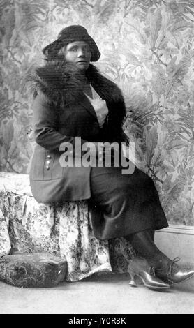 Sitzendes Porträt, eine reife afroamerikanische Frau, trägt dunkles Kleid, Mantel, Pelztuch und Hut, florale Tapete im Hintergrund, ernster Ausdruck, 1920. Stockfoto