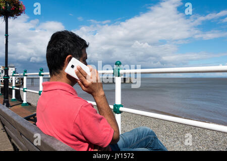 Im mittleren Alter indischer Mann sitzen auf einer Bank auf der Promenade in der Nähe von Llanberis Pier mit Blick auf das Meer über sein Smartphone. Stockfoto