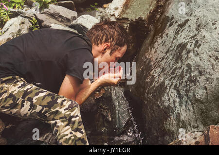 Junge Wanderer Trinkwasser Strom Wasser in Berg. Die durstige Tourist eifrig Getränke Wasser aus einem Wasserfall an einem Gebirgsfluss. Stockfoto