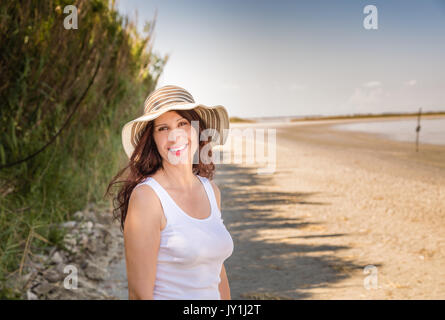 Schöne reife vollbusige Frau in sehr guter Form mit breiten Hut lächelnd auf einem Sun split Strand Stockfoto