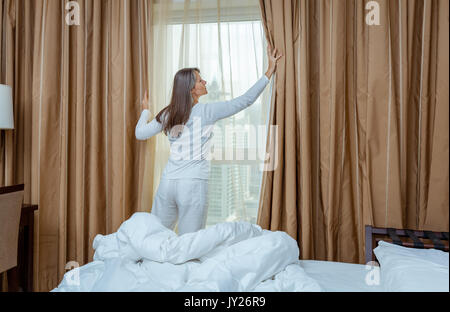 Reife Frau ist in ihrem Schlafzimmer in einem Hotel/Apartment Vorhänge öffnen. Stockfoto