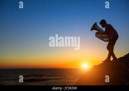 Silhouette des Musikers mit Tuba auf felsigen Küste bei Sonnenuntergang. Stockfoto