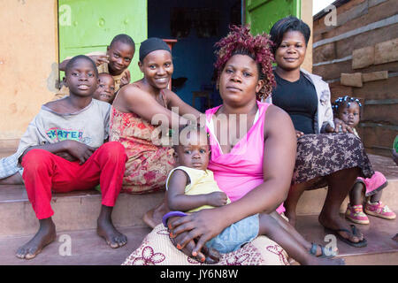 Lugazi, Uganda. 13. Juni 2017. Eine Gruppe von lächelnden afrikanischen Frauen und Kinder sitzen zusammen auf der Treppe vor einer ländlichen Shop. Stockfoto