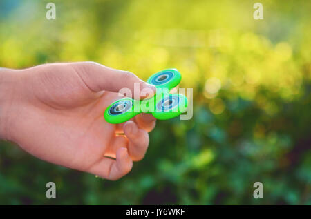 Junger Mann hand mit trendigen antistress Gadget zappeln Spinner. Mann spielt mit grünen Spinner im Freien auf der hellen Bokeh. Flache Freiheitsgrad. Stockfoto