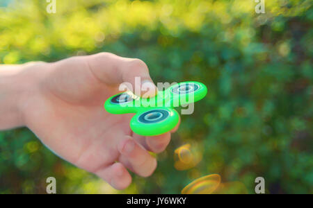 Junger Mann Hand beliebte antistress Gadget zappeln Spinner. Mann spielt mit grünen Spinner im Freien bei Sonnenuntergang Strahlen auf der hellen Bokeh. Shallo Stockfoto