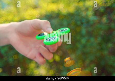 Teenager Hand antistress zappeln Spinner. Junge spielt mit grünen Spinner im Freien bei Sonnenuntergang Strahlen auf die bunten Bokeh. Flache Freiheitsgrad. Stockfoto