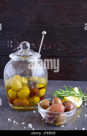 Grüne und schwarze Oliven in Olivenöl in einer traditionellen Glasschale mit speziellen Löffel mit ein paar Oliven in einem kleinen Glasgefäß mit Meersalz, Zitrone, Rosmarin auf einem dunklen Hintergrund Stockfoto
