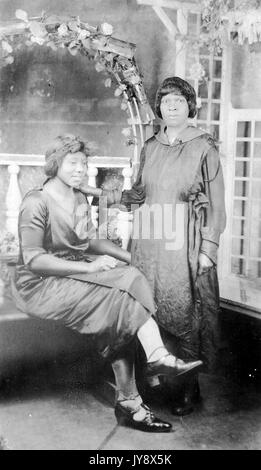 In voller Länge Porträt von zwei Afrikanische amerikanische Frauen, die Frau auf der linken Seite sitzen, mit einem grinsenden Gesichtsausdruck und gekreuzten Beinen, die andere Frau, die ihr mit der einen Hand auf die Schulter der sitzende Frau, mit einem überraschten Gesichtsausdruck, beide Frauen tragen dunkle Kleider, 1920. Stockfoto