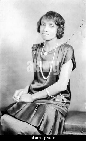 Portrait einer afrikanischen amerikanischen Frau sitzt mit ihr die Hände in ihrem Schoß gefaltet, trug ein Kleid und eine Perle Halskette, 1915. Stockfoto