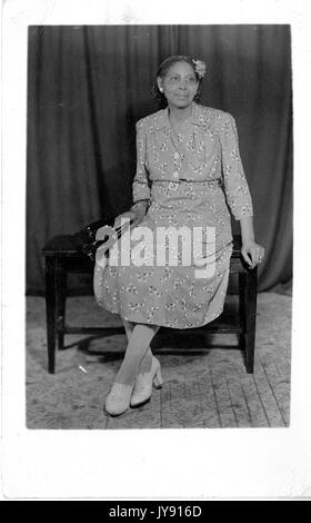 In voller Länge Porträt der afroamerikanischen Frau sitzt auf einem Holztisch, trägt ein gemustertes Kleid und weiße Schuhe und eine schwarze Handtasche, 1920. Stockfoto