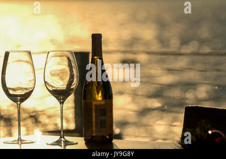 Zwei leere Weingläser mit einer Flasche am Strand home Tabelle mit späten Tag Sonne Beleuchtung das Wasser im Hintergrund. Stockfoto