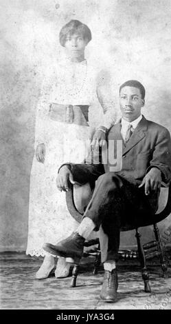 Junge afrikanische - amerikanisches Paar für ein Portrait Foto posieren, der Mann, der auf einem Stuhl sitzend, die Frau hinter ihm stand und tragen weiße Kleid, 1932. Stockfoto