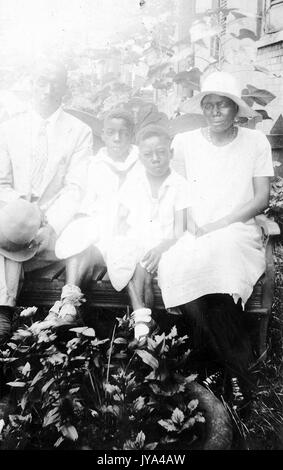 Familie der afrikanisch-amerikanischen Landarbeiter, auf einer Bank in einer Umgebung mit Pflanzen im Hintergrund sitzt ein Mann und eine Frau mit zwei Jungs, der Mann mit dem Hut in der Hand und die Frau in weißem Kleid, original Foto sehr überbelichtet ist, 1932. Stockfoto