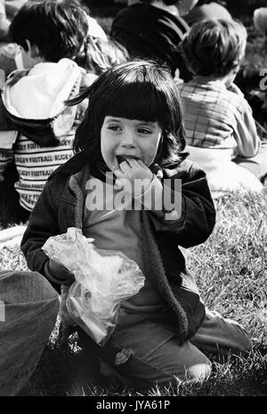 Ein junges Mädchen sitzt auf dem Gras essen Karneval Essen aus einer Plastikhülle, umgeben von anderen kleinen Kindern und Familien im Hintergrund, während 3400 auf Stage Festival an der Johns Hopkins University, Baltimore, Maryland, April 1978. Stockfoto