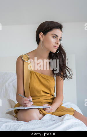 Junge Frau mit langen schwarzen Haaren, auf dem Bett zu sitzen. Sie trägt ein langes braunes Kleid. Stockfoto