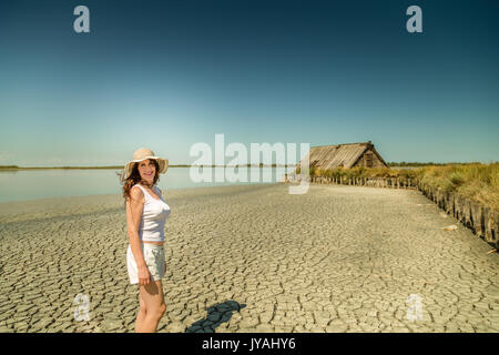 Attraktive reife vollbusige Frau in sehr guter Form mit breiten Hut, stehend auf einem Sun split Strand mit Stroh Hut Stockfoto