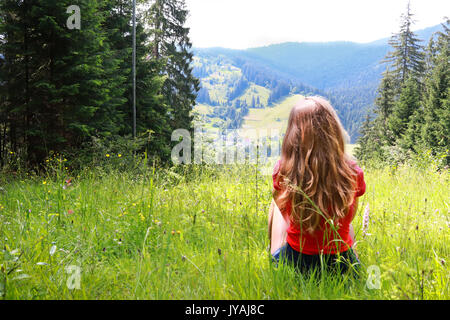 Junge braunhaarige Frau weiblich mit lockigem Haar sitzt am grünen Gras glade am Berg Landschaft Hintergrund im sonnigen Sommertag. In der Ukraine. Karpaten Stockfoto