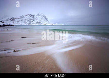 Wellen von der eisigen Meer am Strand im Hintergrund die schneebedeckten Gipfel Pollen Vareid Flakstad Lofoten Inseln Norwegen Europa Stockfoto