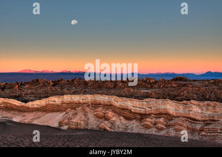 Der Mond scheint über den Gipfeln, die das Tal des Mondes umgeben, durch das Licht des Sonnenuntergangs beleuchtet. Atacama Wüste. Chile. Südamerika Stockfoto