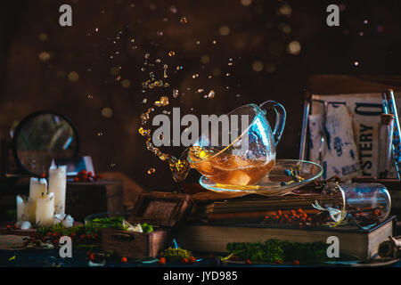 Kaffee Splash in einem Glas Schale auf einem hölzernen Hintergrund mit Kerzen, Geheimnis, Zeitungsausschnitte, Bücher, Laub und Moos. Aktion noch Leben mit Tee Tropfen bokeh Stockfoto