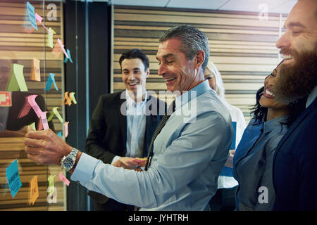 Seitenansicht der lächelnden Geschäftsleute berühren und auf Haftnotizen auf Glas im Büro Stockfoto