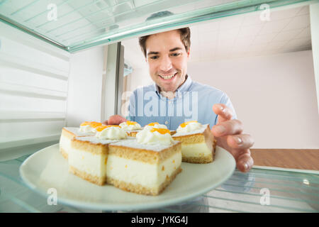 Porträt der jungen gutaussehenden Mann unter Kuchen Blick aus dem Kühlschrank Stockfoto