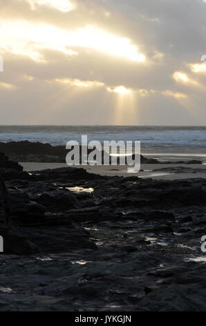 Ein Wolkenbruch Sonnenuntergang über polzeath Strand in North Cornwall in der Nähe des Camel Mündung Lichtstrahlen, die von den Wolken über dem Meer und Surfer Stockfoto