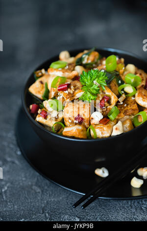 Rühren gebratener Tofu mit Cashew, Chili und Zwiebeln in schwarz Schüssel auf dunklem Hintergrund. Asiatische chinesische Küche Konzept Stockfoto