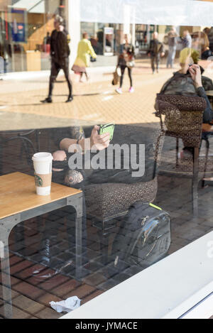 Sitzende Frau verwendet ein Handy im Starbucks mit ihrem Gesicht und Oberkörper von einem Glas Fenster in Richtung der High Street mit Menschen zu Fuß wider Stockfoto