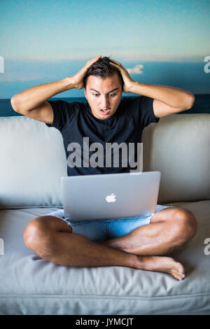 Schockiert, überrascht Mann am Laptop überrascht und mit offenen Mund und großen Augen überrascht. Lustig junge männliche Modell draußen sitzen auf dem Sofa. Stockfoto