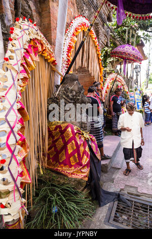 Balinesischen Mann in traditioneller Kleidung, auf ein Handy suchen während einer Königlichen Einäscherung in Ubud, Bali. Bunten Dekorationen auf der linken Seite. Vertikale Format. Stockfoto